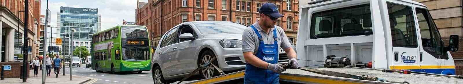 Recovery worker in blue overalls carefully securing a silver car onto a white flatbed truck on a busy Birmingham city street.