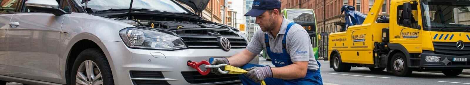 Recovery worker attaching a yellow tow strap to the front bumper of a silver car on a Birmingham street.
